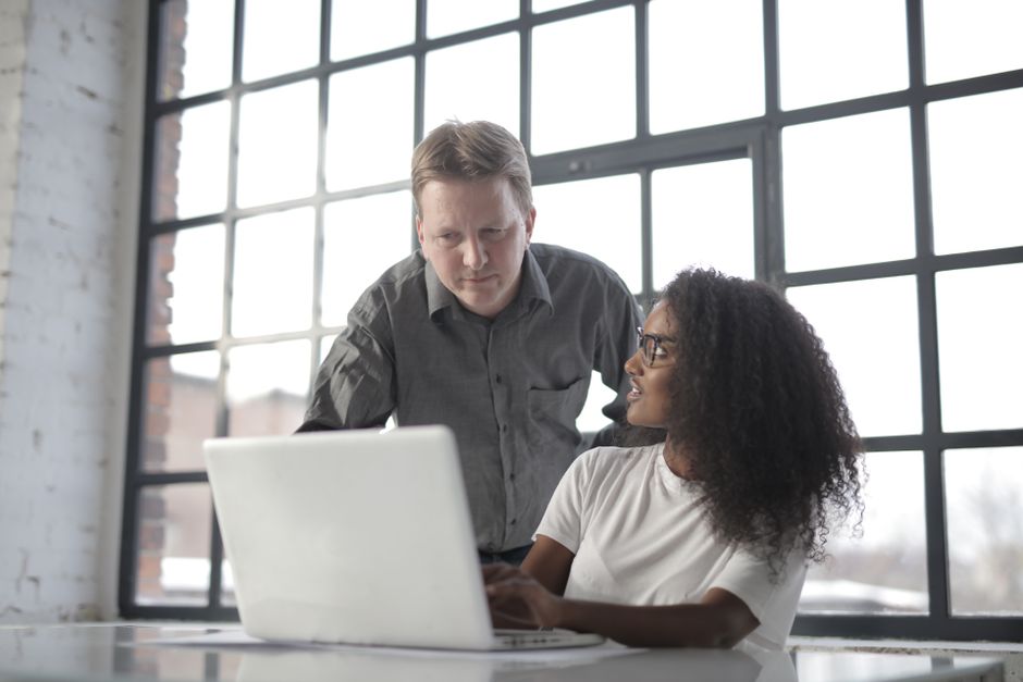 Entrepreneur adulte consultant un ordinateur portable avec une collègue afro-américaine et discutant d'un projet de startup dans un bureau près d'une grande fenêtre.
