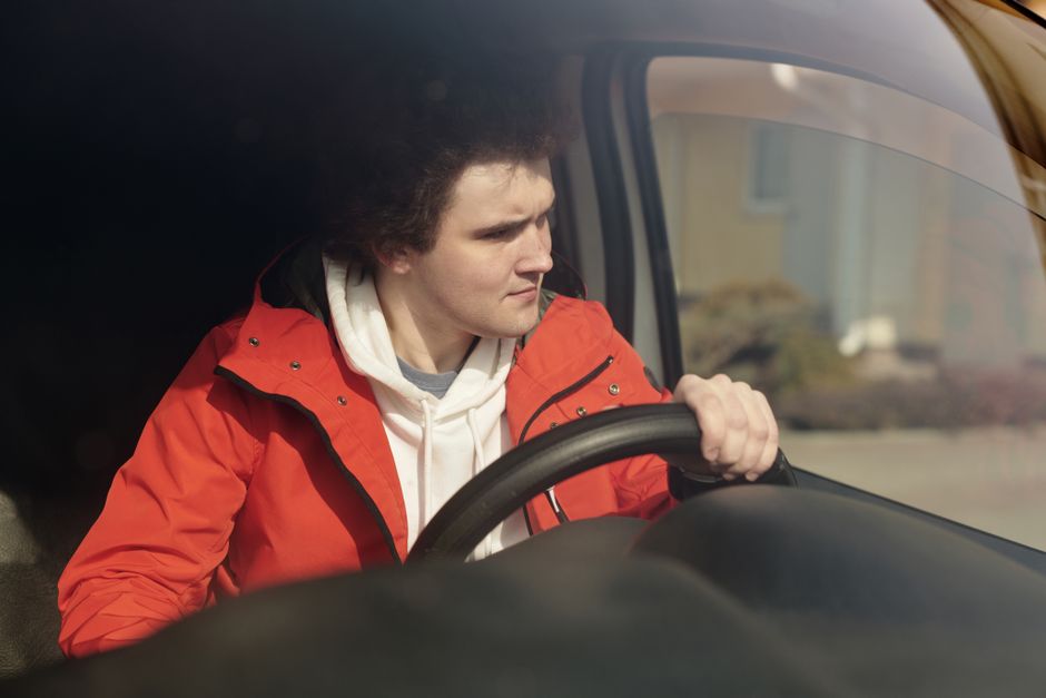 Jeune homme en veste rouge conduisant une voiture avec concentration, profitant d'un trajet paisible.