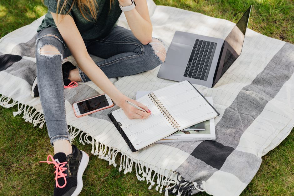 Vue de dessus d'une étudiante anonyme en tenue décontractée écrivant dans un agenda, assise sur une couverture chaude avec un smartphone et un ordinateur portable sur la pelouse d'un parc.