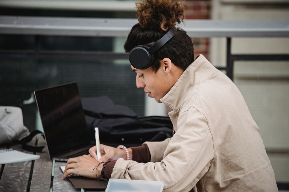 Vue de profil d'un homme d'origine ethnique à la coiffure tendance écrivant dans un carnet en étudiant à une table avec un netbook dans la rue.