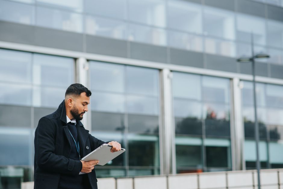 Jeune employé de bureau d'origine ethnique, non rasé, regardant l'heure sur son ordinateur portable devant un bâtiment moderne en ville.