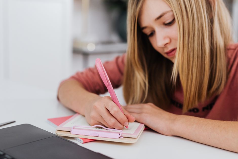 A focused young woman writing in a notebook indoors, showcasing creativity and study habits.
