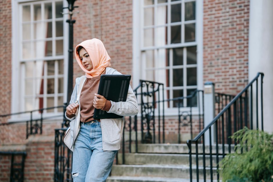 Jeune femme musulmane pensive, en tenue décontractée et foulard traditionnel, descendant les escaliers d'un grand bâtiment dans la rue avec un ordinateur portable.