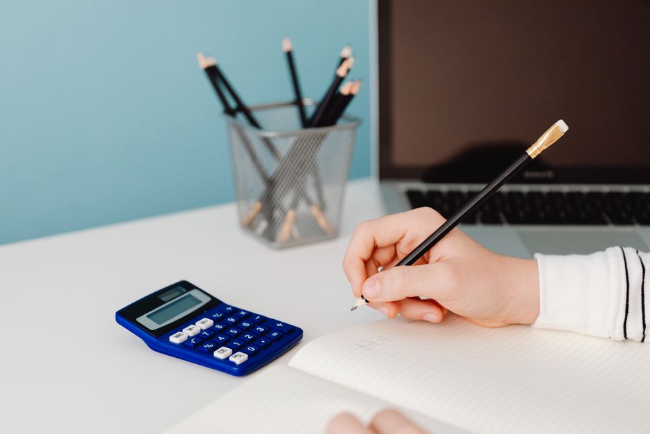 Gros plan d'une main écrivant dans un carnet à côté d'une calculatrice et d'un ordinateur portable sur un bureau.