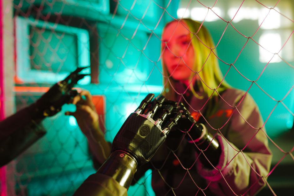 Une femme interagit avec une main robotique à travers une clôture sous des néons.