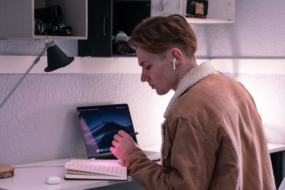 Vue de profil d'un jeune étudiant sérieux portant des écouteurs sans fil, écrivant dans un agenda tout en étant assis à un bureau avec un ordinateur portable.