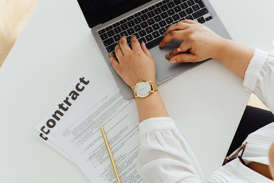 Femme utilisant un ordinateur portable à côté d'un contrat sur un bureau, mettant en avant des thèmes juridiques et professionnels.