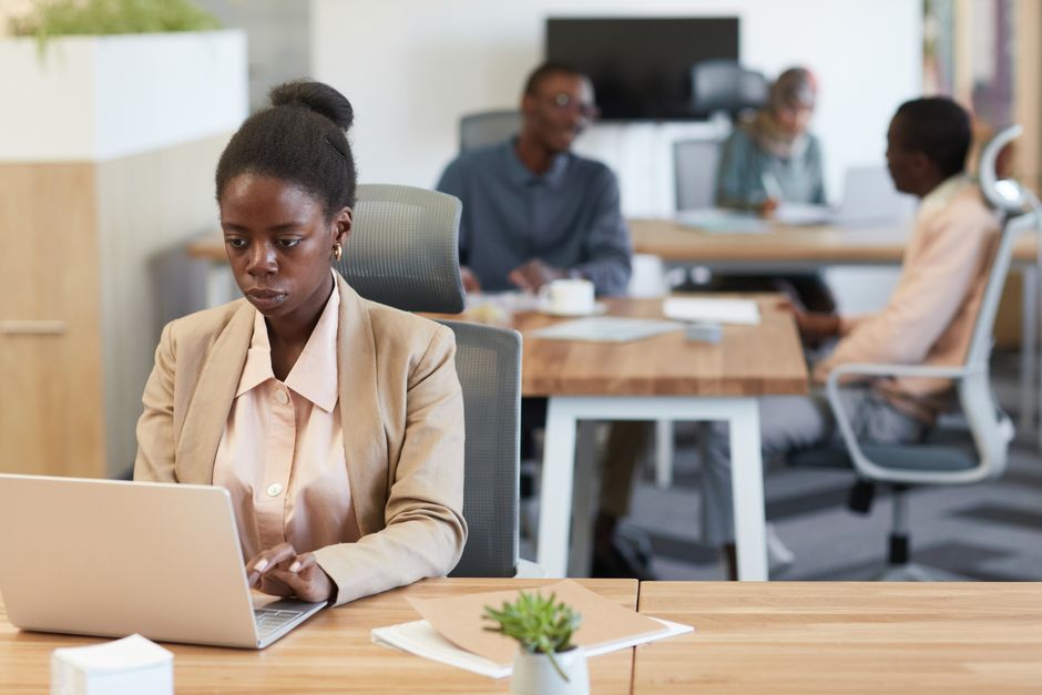 Jeune femme africaine en blazer travaillant avec concentration sur un ordinateur portable dans un bureau animé.