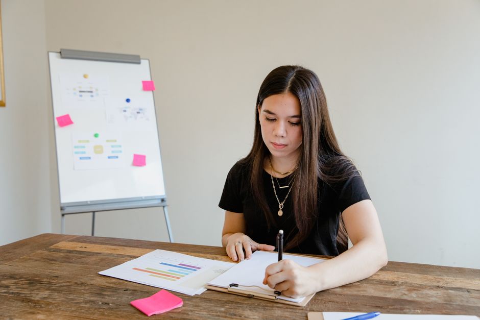 Jeune femme écrivant à une table en bois dans un bureau avec un tableau blanc en arrière-plan.